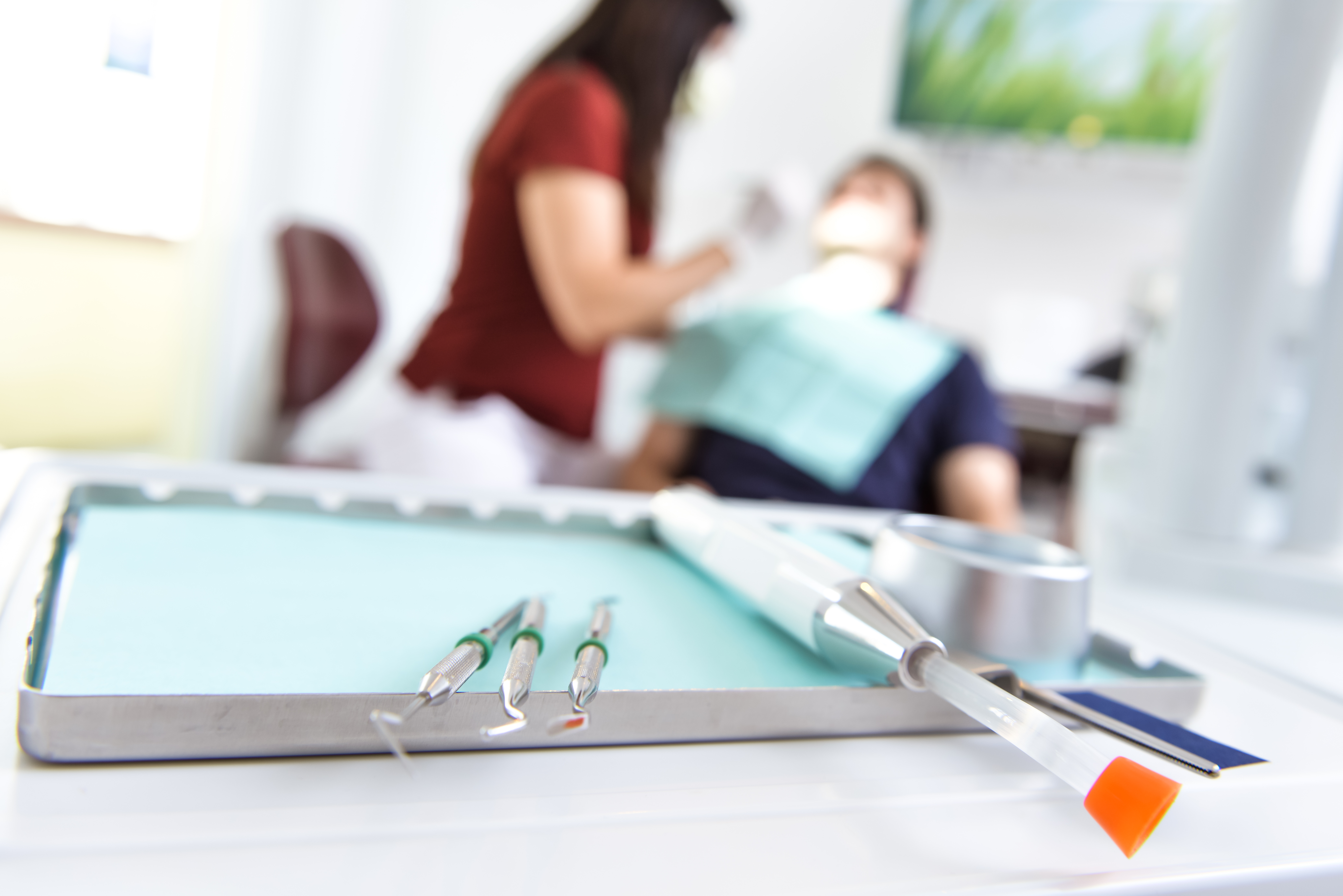 Close-up of dental instruments arranged on a metal tray with a blurred dentist and patient in the background. Dr. Anne Kehler Zahnarzt Berlin Lichtenberg